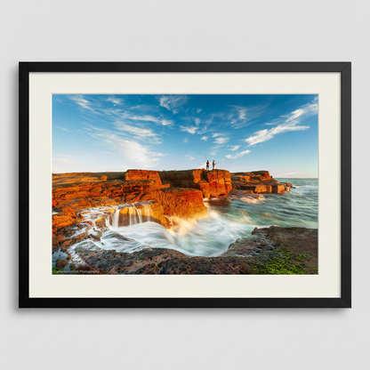 A landscape photograph featuring two figures fishing from a cliff edge with a vibrant sunset in the background, over a body of water. Norah Head lighthouse, NSW coast — fine art coastal wall art
