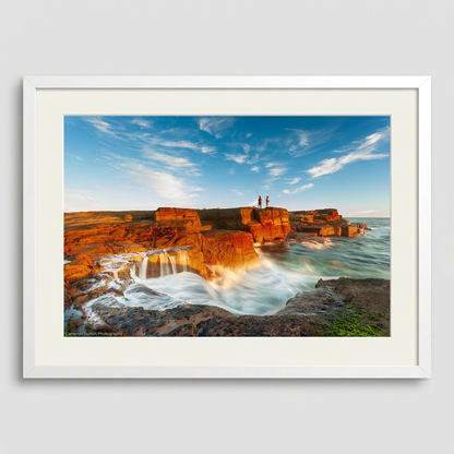 A landscape photograph featuring two figures fishing from a cliff edge with a vibrant sunset in the background, over a body of water. Norah Head lighthouse, NSW coast — fine art coastal wall art