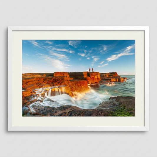 A landscape photograph featuring two figures fishing from a cliff edge with a vibrant sunset in the background, over a body of water. Norah Head lighthouse, NSW coast — fine art coastal wall art