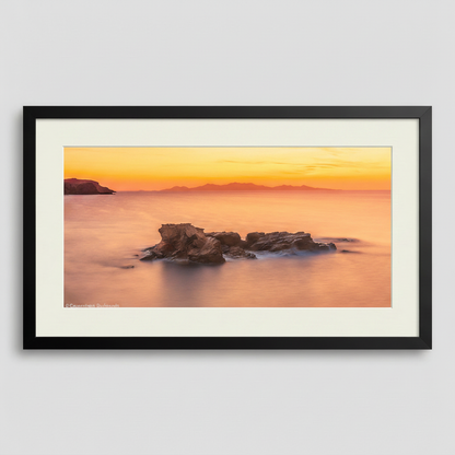 A photograph depicting a sunset over the Aegean Sea from Antiparos, Greece, with a rock formation in the foreground.