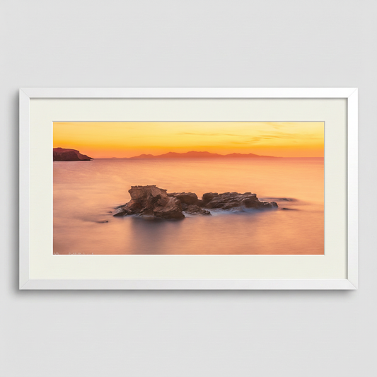 A photograph depicting a sunset over the Aegean Sea from Antiparos, Greece, with a rock formation in the foreground.
