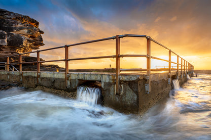 A photography print showing an ocean pool at North Curl Curl during sunrise, featuring rocks, water, and a wooden walkway with a railing.