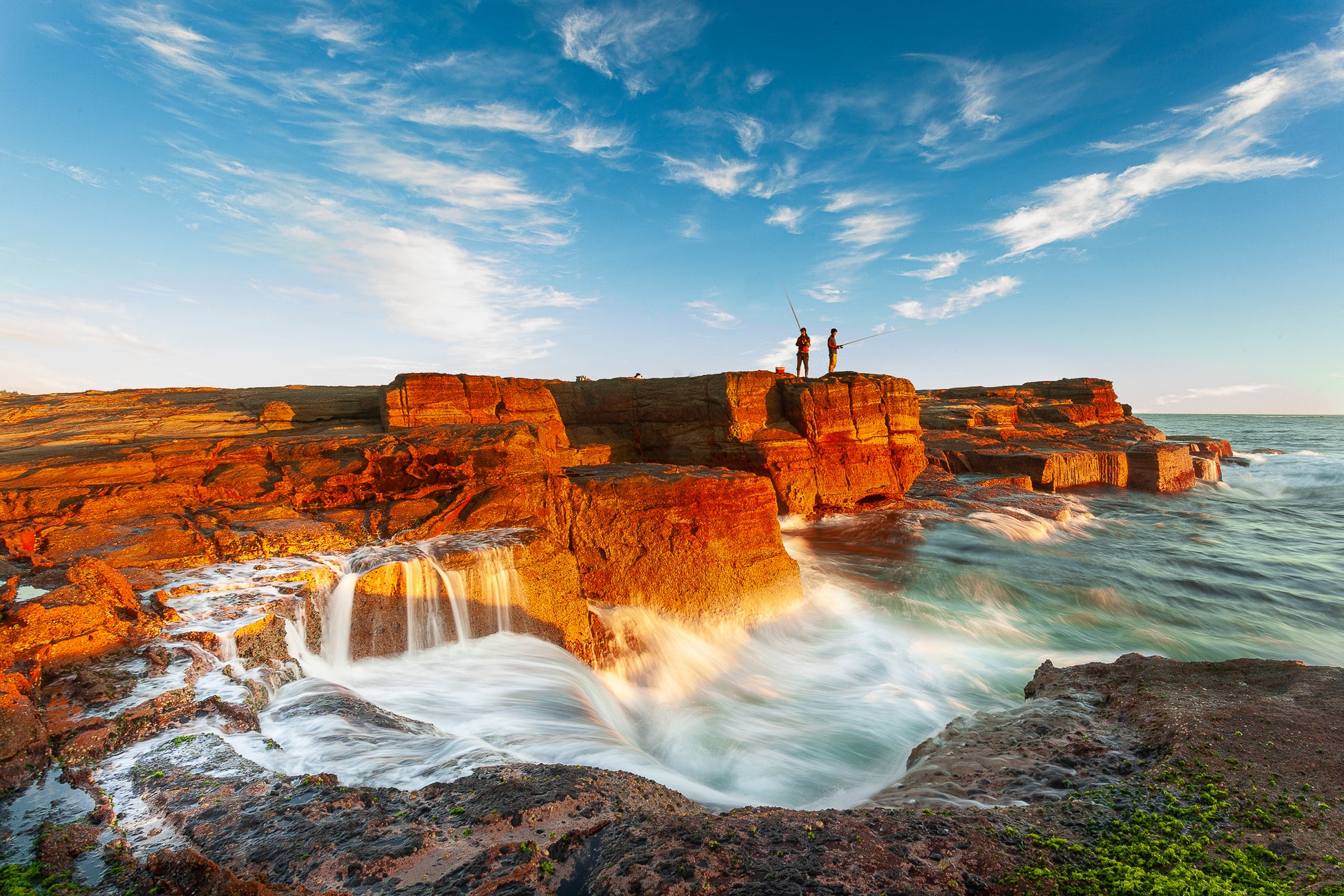 A landscape photograph featuring two figures fishing from a cliff edge with a vibrant sunset in the background, over a body of water. Norah Head lighthouse, NSW coast — fine art coastal wall art