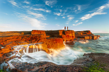 A landscape photograph featuring two figures fishing from a cliff edge with a vibrant sunset in the background, over a body of water. Norah Head lighthouse, NSW coast — fine art coastal wall art